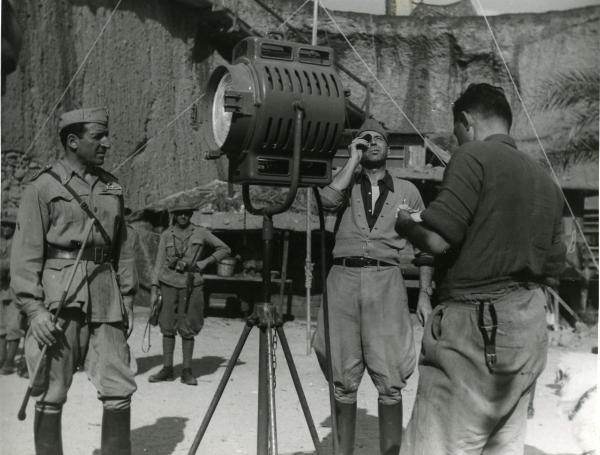 Fotografia sul set di "Giarabub" - Alessandrini, Goffredo, 1942 - A sinistra, Carlo Ninchi guarda verso un attore non identificato con un binocolo in mano. A destra, un operatore non identificato di spalle. I tre sono attorno a un faro.