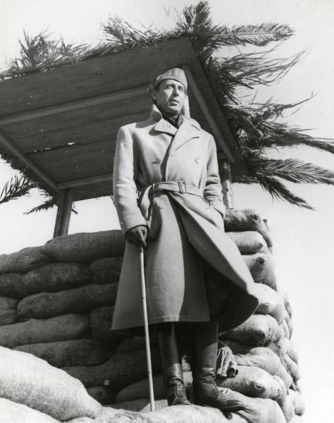 Fotografia sul set di "Giarabub" - Alessandrini, Goffredo, 1942 - Vicino a un fortino fatti di sacchi: figura intera del regista Goffredo Alessandrini in cappotto mentre tiene in mano un bastone.