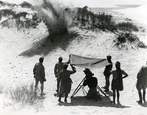 Fotografia sul set di "Giarabub" - Alessandrini, Goffredo, 1942 - Alcuni operatori reggono un telo sopra l'operatore con la cinepresa. L'operatore di camera sta riprendendo un'esplosione in mezzo al deserto.