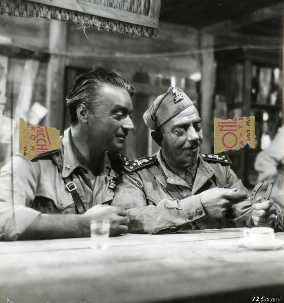 Scena del film "Giarabub" - Alessandrini, Goffredo, 1942 - Bruno Smith, a sinistra, e Corrado De Cenzo, a destra, entrambi appoggiati al bancone di un bar, guardano un ferro di cavallo che tiene in mano De Cenzo.