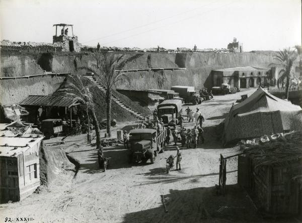 Scena del film "Giarabub" - Alessandrini, Goffredo, 1942 - Alcuni mezzi militari con sopra soldati non identificati stanno passando in mezzo a un vialone di una cittadina.