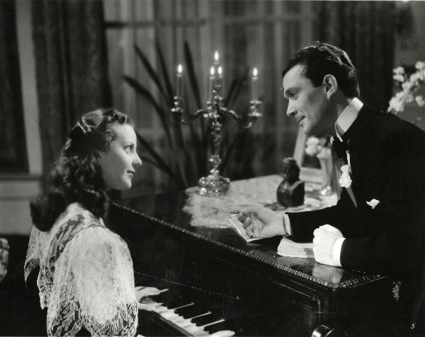 Scena del film "Gioco d'azzardo" - Bassi, Parsifal, 1943 - A sinistra, Lia Orlandini suona il pianoforte. A destra, Maurizio D'Ancora è appoggiato al pianoforte. I due si guardano intensamente e si sorridono.