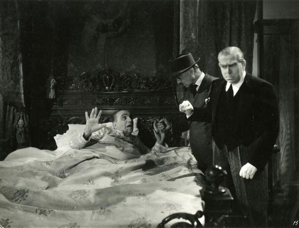 Scena del film "Gioco d'azzardo" - Bassi, Parsifal, 1943 - Vasco Creti, sdraiato in un letto alza le mani in segno di resa poiché, a destra, Arturo Bragaglia lo intima con una pistola. Di fianco a Bragaglia, un attore non identificato.