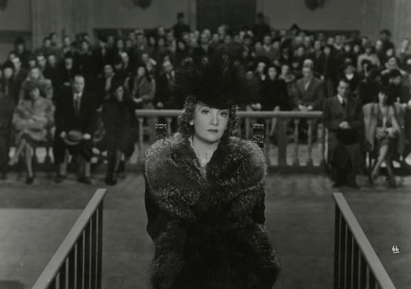 Scena del film "Gioco pericoloso" - Malasomma, Nunzio, 1942 - In un'aula di tribunale: in primo piano Elsa Merlini guarda dritto di fronte a sé. In secondo piano, molti attori non identificati sono seduti e guardano la Merlini.