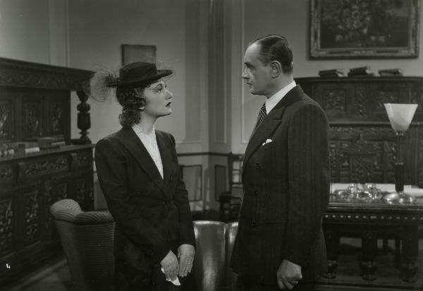 Scena del film "Gioco pericoloso" - Malasomma, Nunzio, 1942 - In uno studio: a sinistra, Elsa Merlini, tenendo tra le mani una piccola borsa, guarda a destra Guido Notari che ricambia lo sguardo.