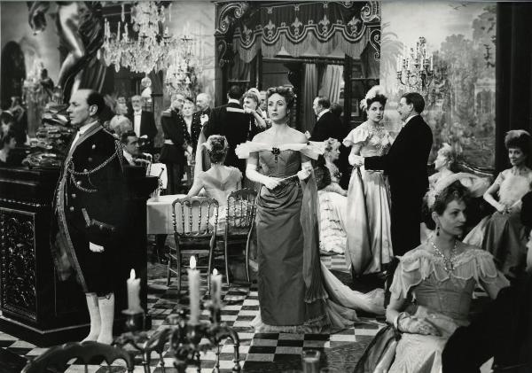 Scena del film "I gioielli di Madame de..." - Ophuls, Max, 1953 - In una sala adibita a festa, al centro, Danielle Darreaux cammina fiera con un ventaglio chiuso nelle mani. Attorno, molti attori non identificati danzano e parlano fra loro.