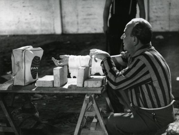 Scena del film "I giorni contati" - Petri, Elio, 1962 - Salvo Randone mentre è seduto di fronte un tavolo in legno con dei mattoni su cui appoggia il braccio destro fasciato.