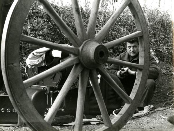 Fotografia sul set di "Un giorno da leoni" - Loy, Nanni, 1961 - Dietro la ruota di un carro, il regista Nanni Loy seduto per terra con di fianco delle attrezzature per la ripresa.