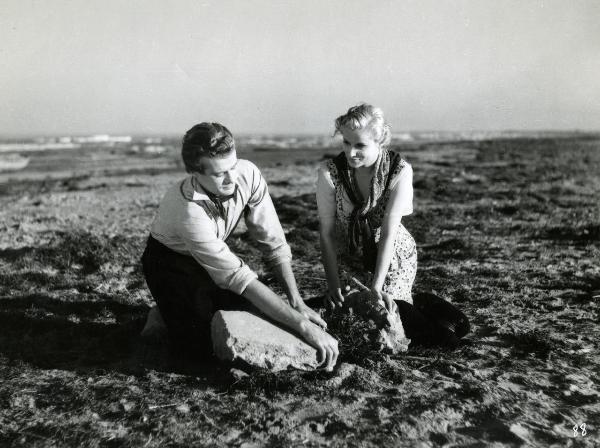 Scena del film "Giorno di festa" - Randone, Belisario, De Ribon, Roberto, 1942 - In riva al mare, Maria Mercander, appoggiandosi a un masso, guarda sorridendo Massimo Serato intento a spostare anch'egli un masso.