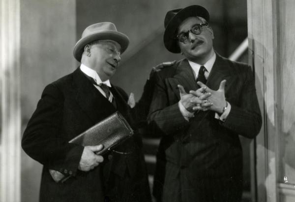 Scena del film "Giorno di nozze" - Matarazzo, Raffaello, 1942 - A sinistra, Carlo Campanini mentre regge sotto il braccio una borsa e poggia la mano sinistra sulla spalla di un attore non identificato che tiene le mani incrociate.