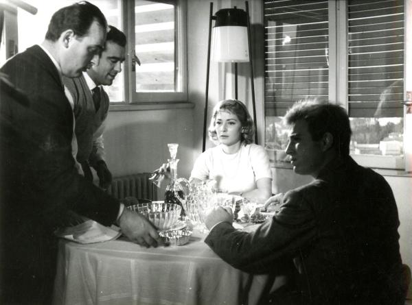 Fotografia sul set di "Giovani mariti" - Bolognini, Mauro, 1957 - Seduti a tavola, l'attrice Rosy Mazzacurati e un attore non identificato. In piedi, mentre sistemano gli oggetti sulla tavola, il regista Mauro Bolognini e Marcello Gatti.