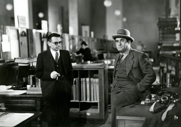 Scena del film "Gioventù perduta" - Germi, Pietro, 1947 - A destra, con una gamba su una scrivania, Nando Bruno con le mani dentro le tasche dei pantaloni. A sinistra, un attore non identificato mentre guarda Nando Bruno.