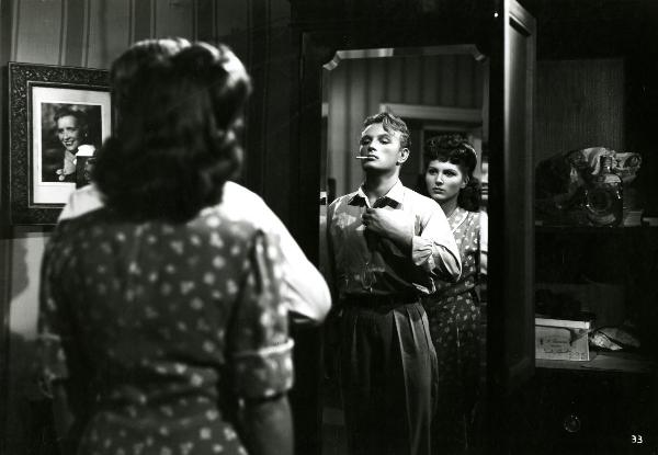 Scena del film "Gioventù perduta" - Germi, Pietro, 1947 - Riflessi in uno specchio, Jacques Sernas e Carla Del Poggio. Con la sigaretta in bocca, l'attore, con la mano destra si abbottona la camicia. L'attrice, dietro di lui, lo guarda.