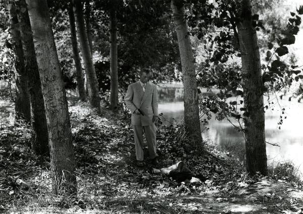Scena del film "Gioventù perduta" - Germi, Pietro, 1947 - In mezzo a degli alberi, sulla riva di un fiume, Jacques Sernas in piedi punta la pistola contro un'attrice non identificata distesa a terra di fronte a lui.