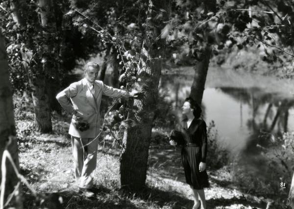 Scena del film "Gioventù perduta" - Germi, Pietro, 1947 - Sulla riva di un fiume, Jacques Sernas appoggiato con la mano sinistra a un albero guarda dritto di fronte a sé. A destra, Carla Del Poggio, con la borsetta sotto braccio, lo guarda.