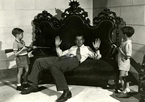 Scena del film "Gioventù perduta" - Germi, Pietro, 1947 - Al centro, semi-sdraiato su un divano intarsiato, Massimo Girotti con le mani in alto. Ai lati, due bambini non identificati gli puntano contro fucile e pistola.