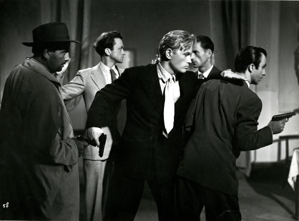 Scena del film "Gioventù perduta" - Germi, Pietro, 1947 - Jacques Sernas tiene la pistola in una mano e poggia il braccio sulle spalle di un attore non identificato. A destra, un attore non identificato in impermeabile punta la pistola contro Sernas.