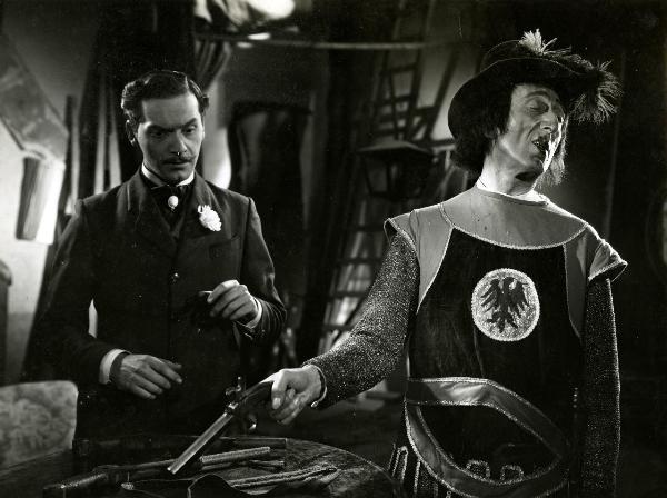 Scena del film "Giù il sipario" - Matarrazzo, Raffaello, 1940 - A destra, Claudio Ermelli in veste da cavaliere, impugna nella mano destra una pistola e rivolge la testa a destra. A sinistra, Andrea Checchi fissa la pistola tenuta in mano da Ermelli.