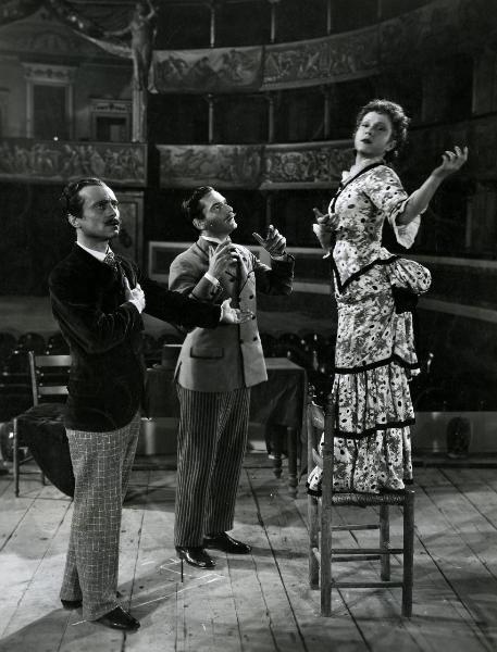 Scena del film "Giù il sipario" - Matarrazzo, Raffaello, 1940 - Sul palco di un teatro, da sinistra a destra: Andrea Checchi, Giulio Panicali e Lilia Silvi mentre gesticolano. L'attrice è in piedi su una sedia.

