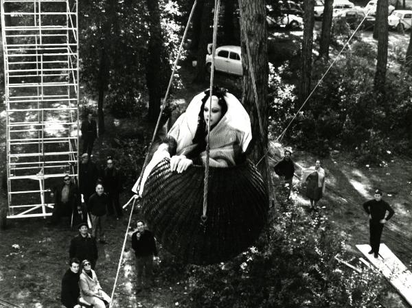 Scena del film "Giulietta degli spiriti" - Fellini, Federico, 1965 - In una cesta sospesa in aria con delle funi, in abiti stravaganti, Sandra Milo. Attorno, operatori non identificati la guardano.




