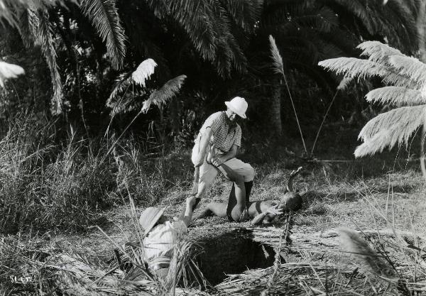 Scena del film "Giungla" - Malasomma, Nunzio, 1942 - Un attore non identificato tiene con la mano sinistra in ostaggio un indigeno mentre con la destra aiuta un altro attore non identificato a uscire da una buca.


