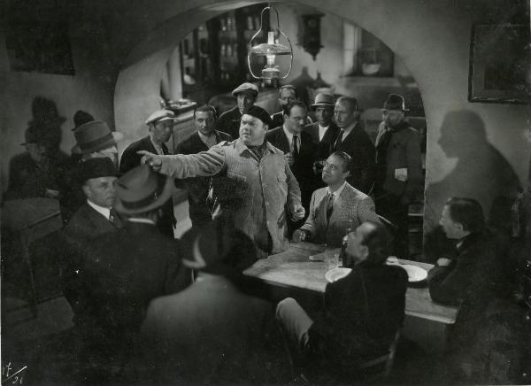 Scena del film "Gran premio" - Musso, Giuseppe, Scarpelli, Umberto, 1957 - Al centro della scena, Juan De Landa indica qualcosa con il braccio destro. Di fianco a lui seduto e sorridente, Bruno Smith lo guarda. Attorno, attori non identificati.



