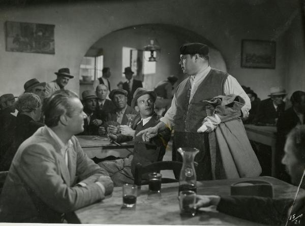 Scena del film "Gran premio" - Musso, Giuseppe, Scarpelli, Umberto, 1957 - In un'osteria: Huan De Landa è in piedi con in mano un cappotto e la mano sinistra fasciata. Subito vicino Bruno Smith. Attorno, attori non identificati.
