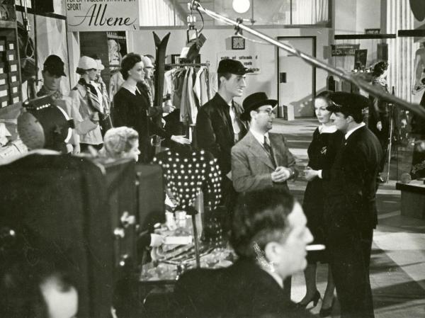 Sul set del film "Grandi magazzini" - Camerini, Mario, 1939 - A destra, il regista Mario Camerini con un cappello in testa spiega a Vittorio De Sica e a Milena Penovich una scena del film.

