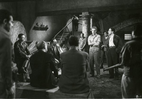 Scena del film "Il grido della terra" - Coletti, Duilio, 1948 - In una sala sotterranea, al centro della scena con le braccia conserte, Renato Bossi. Attorno a lui alcuni attori non identificarti lo guardano.
