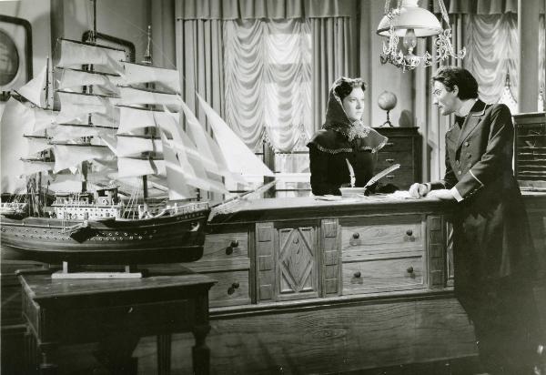 Scena del film "Guarany" - Freda, Riccardo, 1948 - Dietro un bancone, Mariella Lotti guarda un attore non identificato poggiato al banco. A sinistra su un tavolino, la riproduzione in scala di un veliero.
