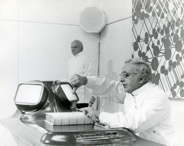 Scena del film "H2S" - Faenza, Roberto, 1971 - In fondo alla stanza, Lionel Stander. In primo piano, un attore non identificato seduto a una scrivania-marchingegno. L'attore tiene il pollice della mano destra rivolto verso il basso.