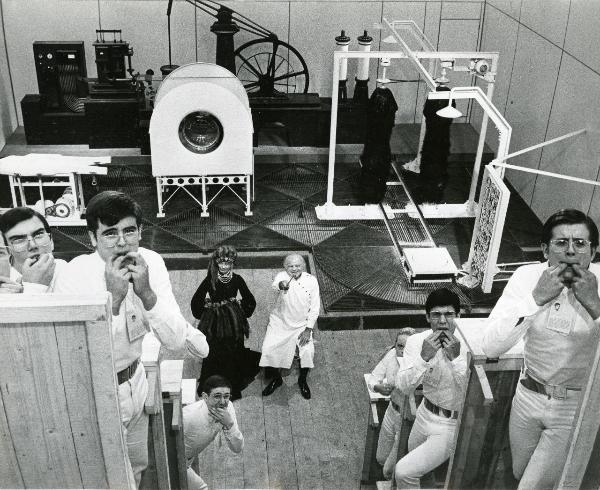 Scena del film "H2S" - Faenza, Roberto, 1971 - Ripresa di un laboratorio pieno di marchingegni. In basso, Lionel Stander, a destra, e Paolo Poli, a sinistra. In primo piano attori non identificati fischiano.
