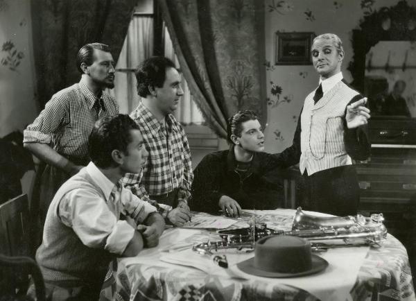 Scena del film "Idillio a Budapest" - Ansoldi, Giorgio, Varriale, 1941 - A un tavolo: Sergio Tofano, a destra, con un gilet a righe  e Michele Riccardini, al lato opposto mentre scrive su un foglio. Attorno, attori non identificati guardano Tofano.