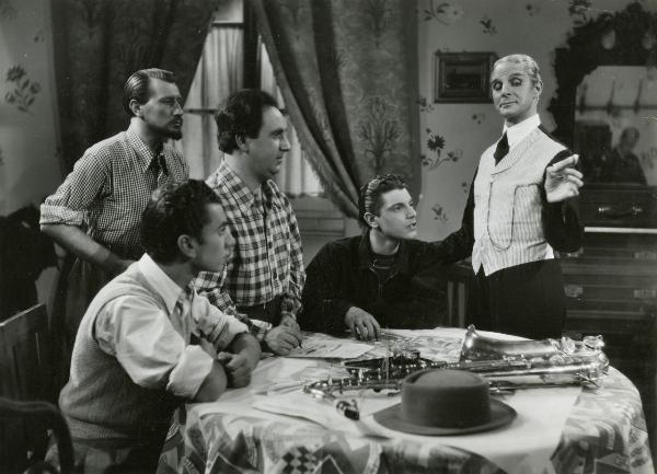 Scena del film "Idillio a Budapest" - Ansoldi, Giorgio, Varriale, 1941 - A un tavolo: Sergio Tofano, a destra, con un gilet a righe  e Michele Riccardini, al lato opposto mentre scrive su un foglio. Attorno, attori non identificati guardano Tofano.