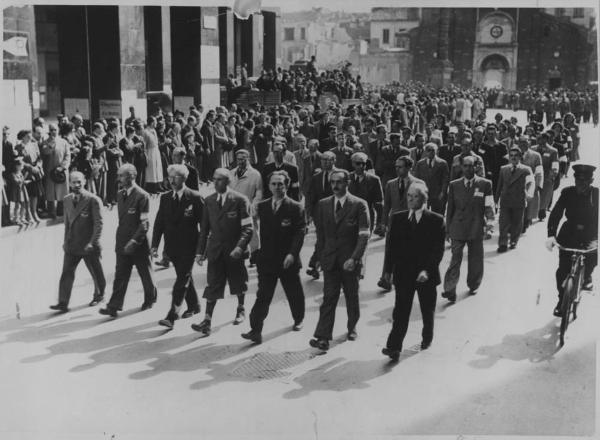 Seconda guerra mondiale - Liberazione - Milano, Piazza San Babila - Sfilata delle Forze di Resistenza - Corteo per le strade della città - Partigiani - In prima fila il comando generale del Corpo Volontari della Libertà (CVL)  - Folla / In prima fila lo Stato Maggiore del Comitato di Liberazione Nazionale Alta Italia (CLNAI), da sx: Mario Argenton, Gian Battista Stucchi, Ferruccio Parri, Raffaele Cadorna, Luigi Longo, Enrico Mattei, Fermo Solari