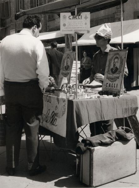 Pescia (PT) - Strada - Mercato - Banco con merce: callifugo Lindangilella - Venditore ambulante con cappello di carta e cliente