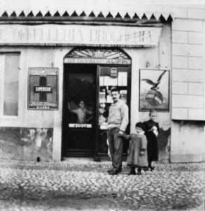 Un negozio in piazza, di fronte al molo. Scritta sulla tenda: "Offelleria Drogheria". Insegne pubblicitarie ai lati della vetrina.