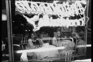 Gruppo di giovani al tavolo di un bar. In primo piano insegna e vetrina del locale.